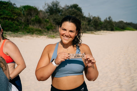 A woman on the beach in activewear smiling at the camera and holding up a Chief Collagen Bar