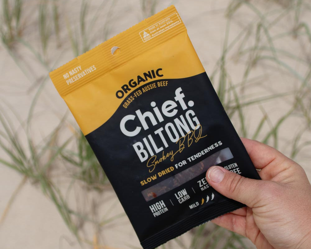 A person's hand holding a bag of Chief Smokey BBQ Biltong against a sand dune backdrop