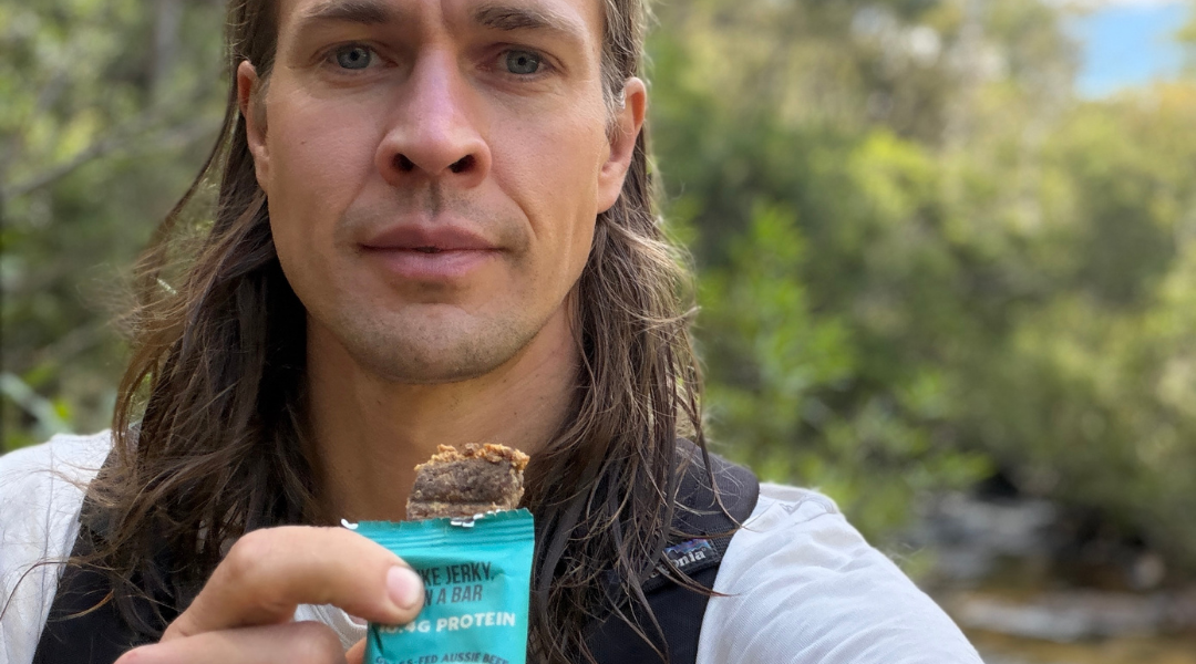 A woman at home in a bright kitchen happily taking a bite of a Chief Collagen Protein bar