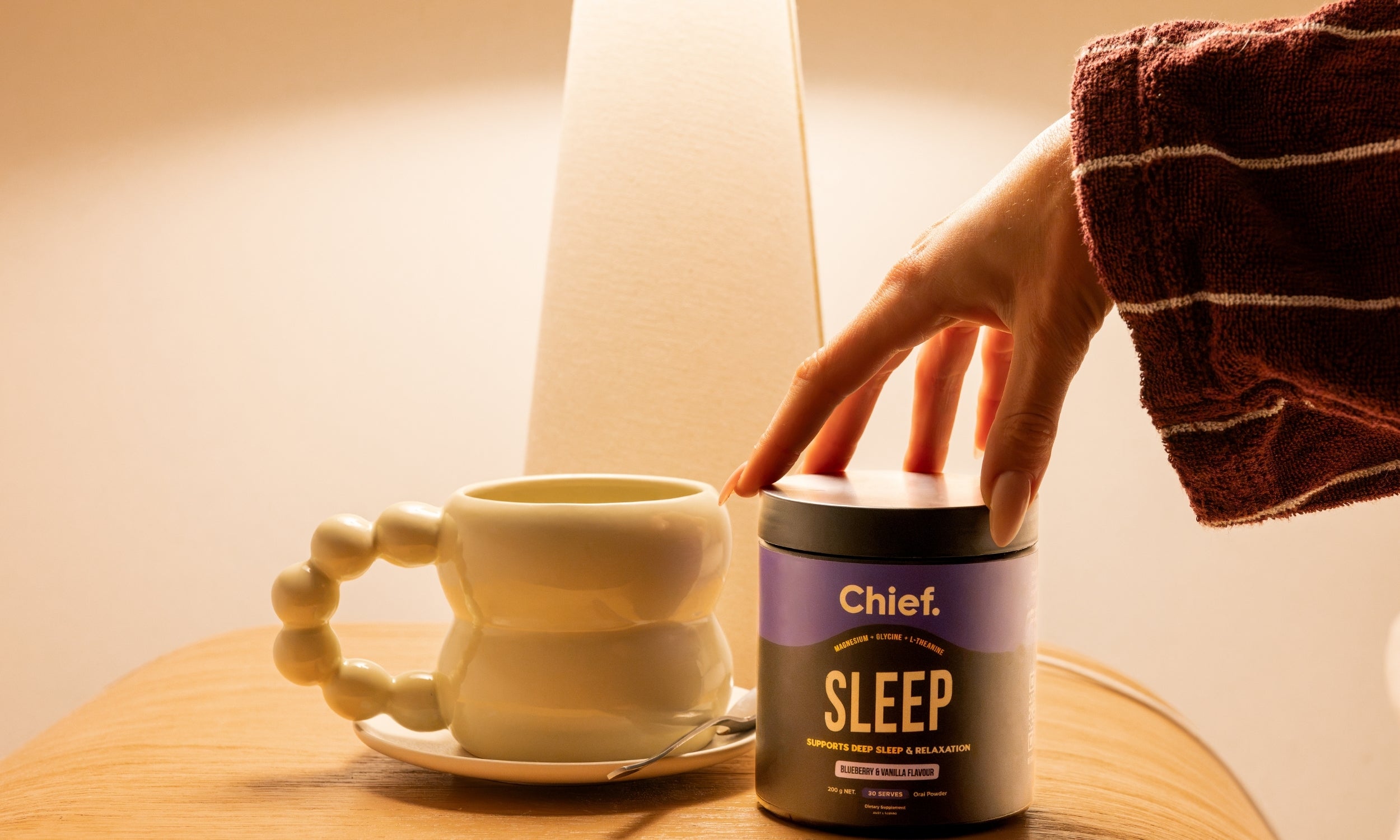 A hand in a striped brown robe reaches for the lid of a dark jar labeled 'Chief. SLEEP' on a wooden table next to a cream-colored ceramic mug under warm, ambient lighting.