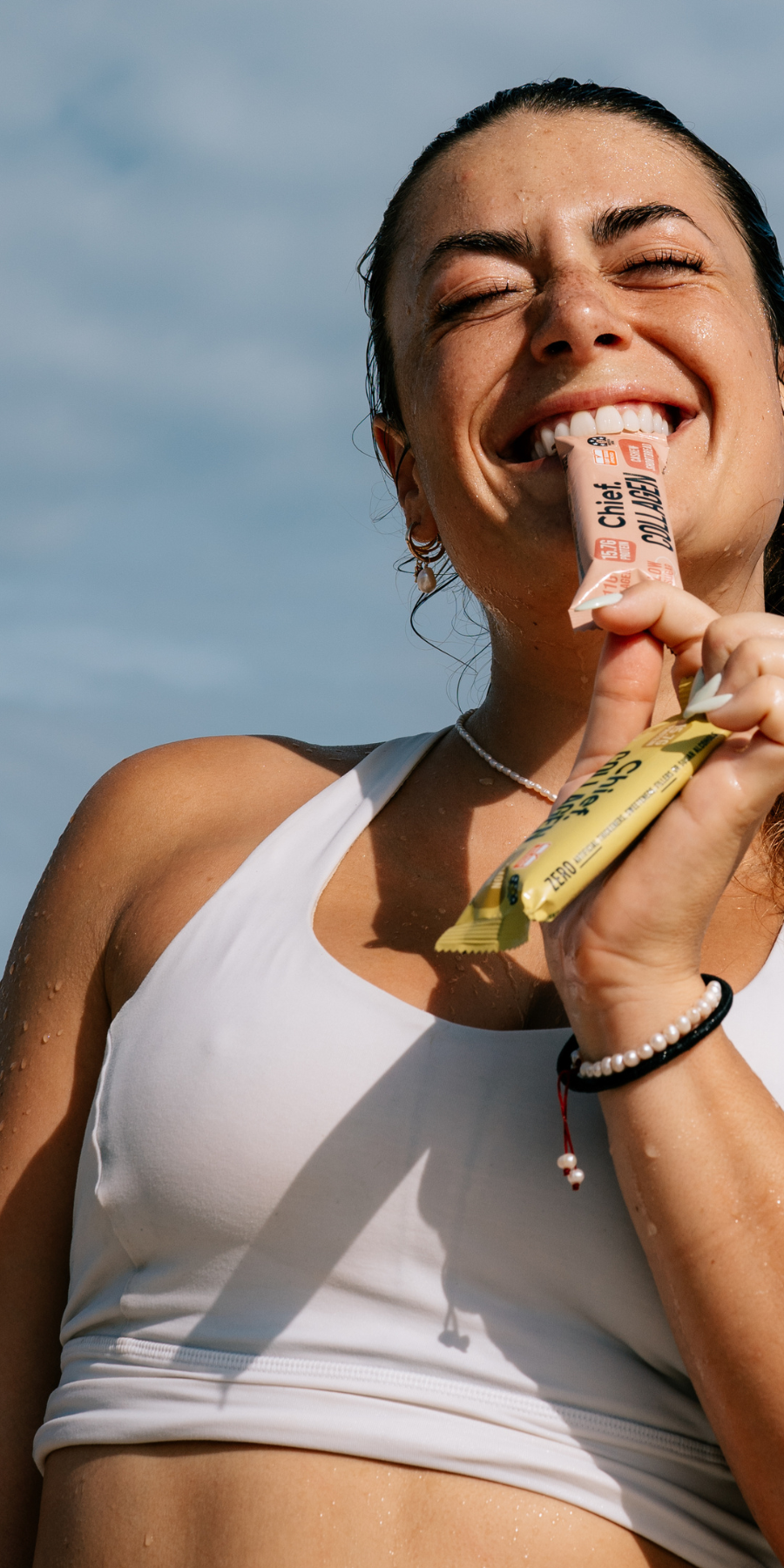 A woman outdoors wearing a white sports bra, smiling as she holds a pink protein powder tube in her teeth and a yellow snack bar in her hand against a blue sky background.